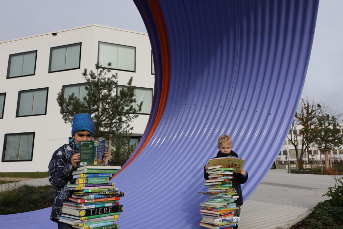 Zwei Jungen stehen hinter zwei Büchertürmen und lesen. Im Hintergrund ist das Fraunhofer Institut IWKS zu sehen