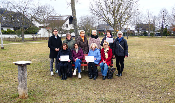 Bild vergr&ouml;&szlig;ern: Auf einer Wiese sitzen vier Frauen neben einander auf der roten Bank. Dahinter stehen sechs weitere Personen. Alle blicken gemeinsam in die Kamera.