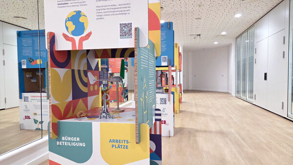 Bild vergr&ouml;&szlig;ern: In einem hellen Raum mit einer verspiegelten Wand stehen hinter einander die vier ringsrum beschrifteten Säulen, aus denen die Wanderausstellung besteht.