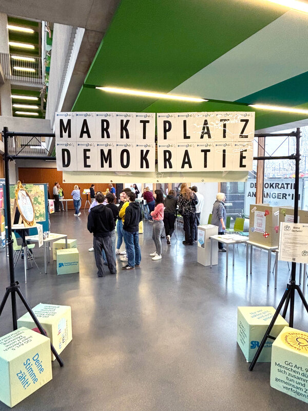 Bild vergr&ouml;&szlig;ern: Unter dem an einer Traverse angebrachten Schriftzug "Marktplatz Demokratie" hindurch ist eine Gruppe junger Menschen zu sehen, die sich an verschiedenen Info-Ständen informieren und besprechen.