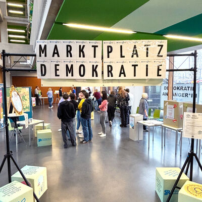 Unter dem an einer Traverse angebrachten Schriftzug "Marktplatz Demokratie" hindurch ist eine Gruppe junger Menschen zu sehen, die sich an verschiedenen Info-Ständen informieren und besprechen.