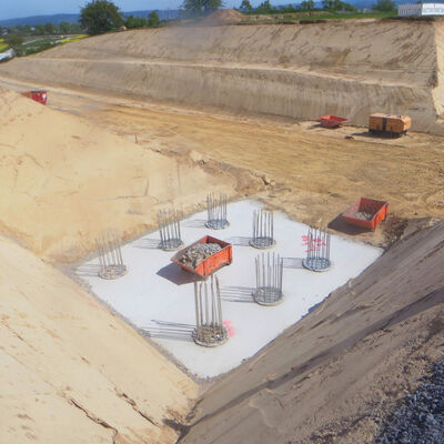 Bild vergr&ouml;&szlig;ern: Abgesenkt zwischen aufegeschütteter Erde sieht man zwei betonierte Fundamente in der Erde, aus denen zahlreiche Stahlstreben nach oben heraus stehen.