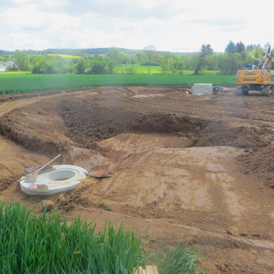 Bild vergr&ouml;&szlig;ern: Am Rande eines weiten Erdlochs sieht man ein betoniertes Bauteil mit einem großen Loch, während auf der anderen Seite des Erdlochs ein Bagger steht.