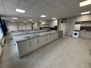 Vier große Küchenzeilen mit grauen Arbeitsflächen und weißen Blenden stehen auf einem grauen Boden, wärend an den Wänden weitere Schränke und Geräte wie eine Waschmaschine und ein Trockner zu sehen sind.