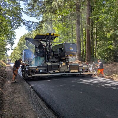 Bild vergr&ouml;&szlig;ern: Eine Baumaschine, so breit wie die Straße selbst, fährt darüber, während auf beiden Seiten jeweils ein Bauarbeiter mit der Maschine interagiert.
