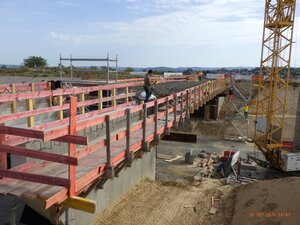 Auf der im Rohbau befindlichen Brücke sind zahlreiche roter Holzbretter als temporäres Geländer verbaut, während man rund um den Rohbau abgetragene und aufgeschüttete Erde sieht sowie einen Kran der rechts neben der Brücke steht