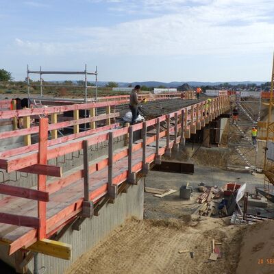 Bild vergr&ouml;&szlig;ern: Auf der im Rohbau befindlichen Brücke sind zahlreiche roter Holzbretter als temporäres Geländer verbaut, während man rund um den Rohbau abgetragene und aufgeschüttete Erde sieht sowie einen Kran der rechts neben der Brücke steht